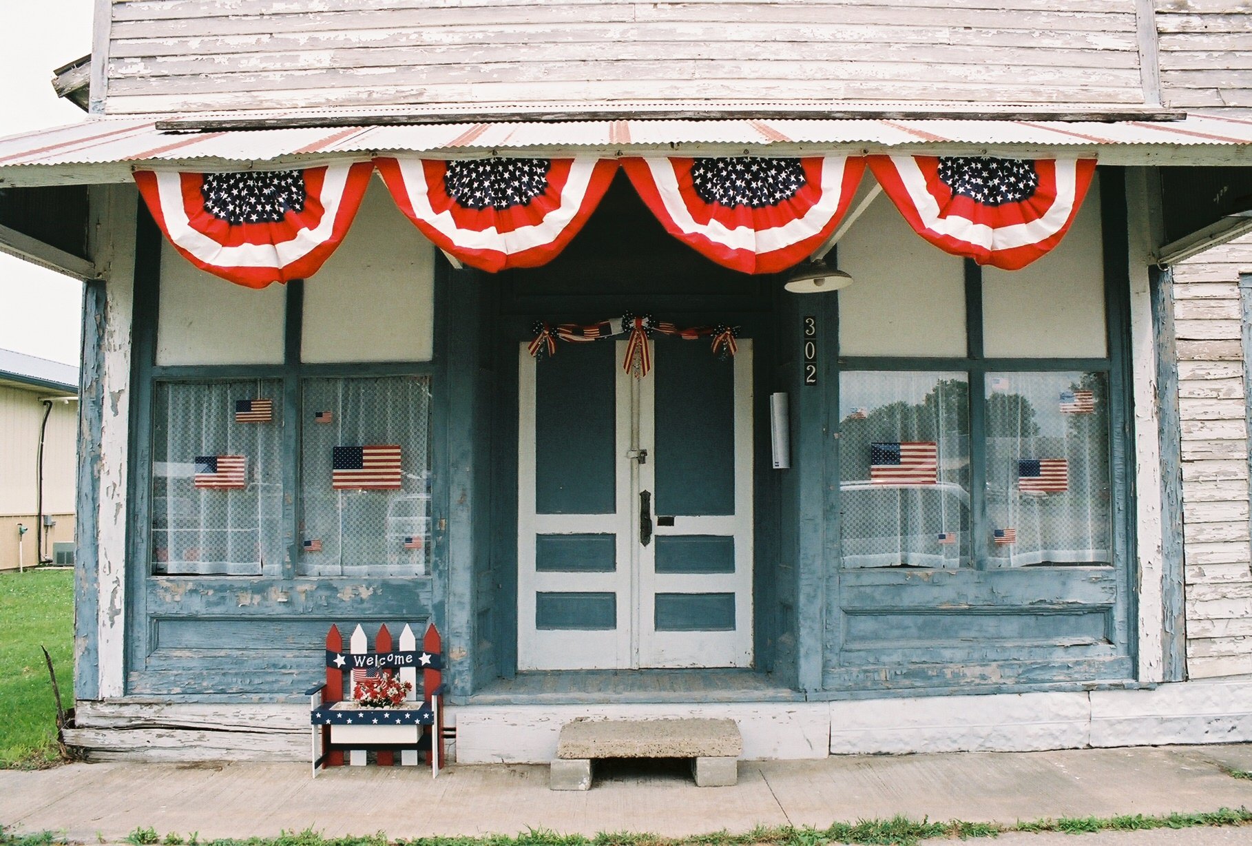 Historic building entrance decorated with red, white, and blue bunting and American flags, featuring a welcoming bench and rustic wooden details. Ideal for showcasing patriotic themes and American heritage.