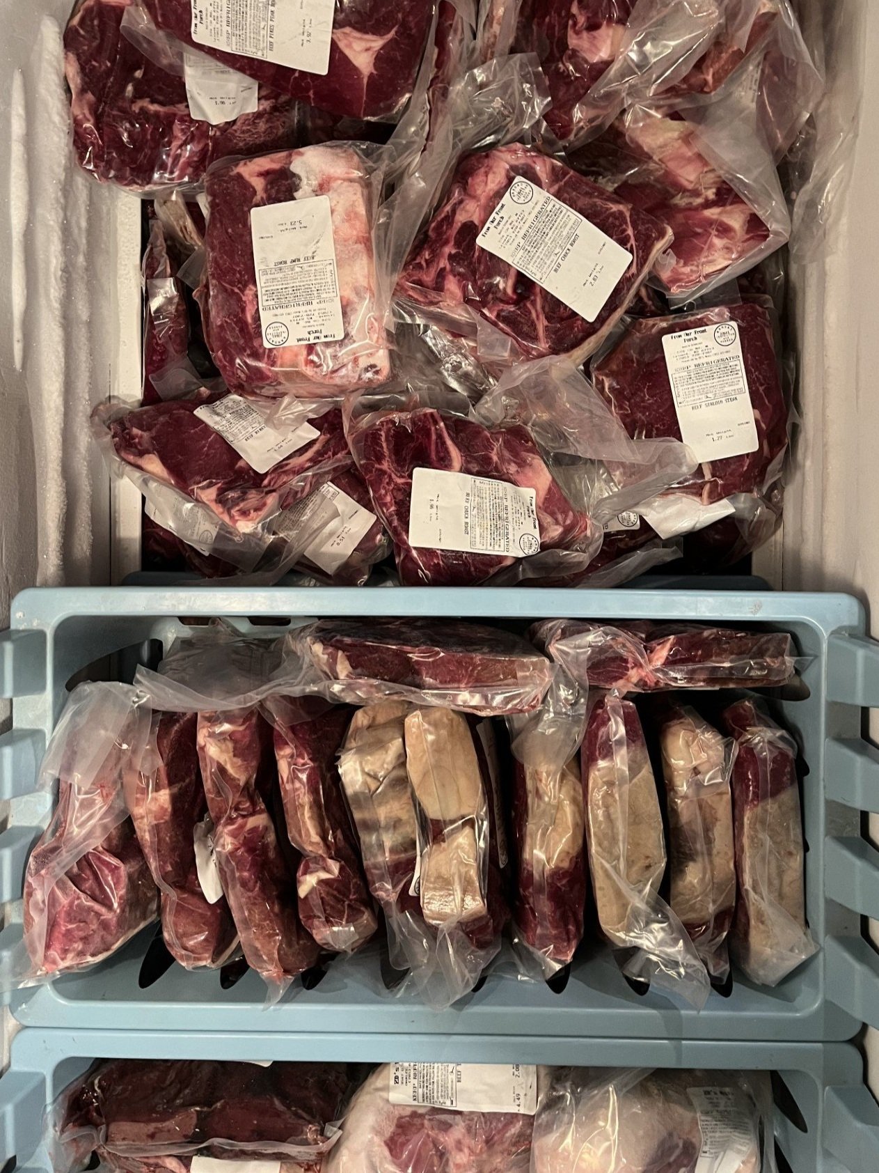 Assorted packages of frozen meat in a cooler, including various cuts of beef, all vacuum-sealed with labels displaying product information.