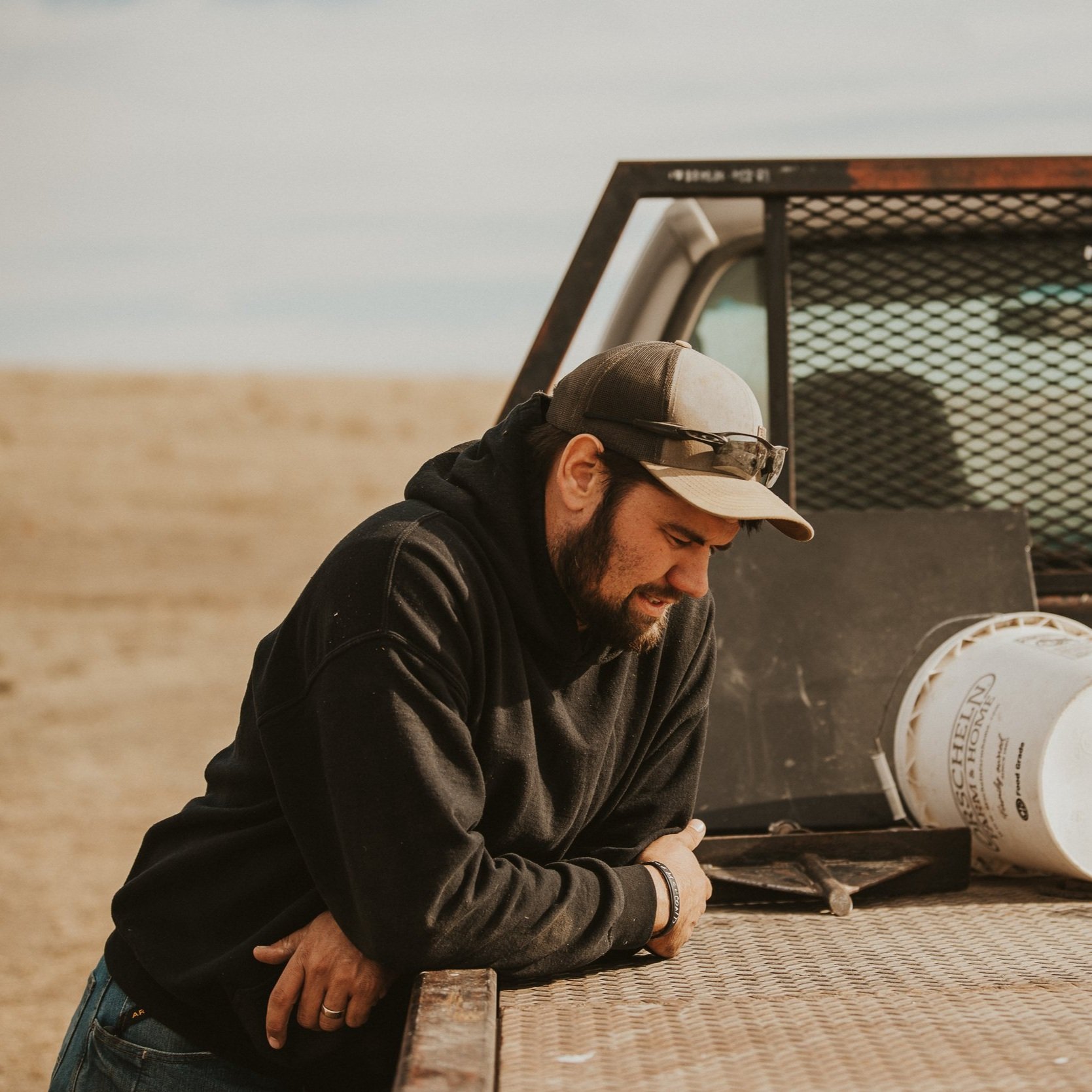Man in a black hoodie and baseball cap leaning over a truck bed, appearing contemplative in an outdoor setting with a blurred landscape in the background.