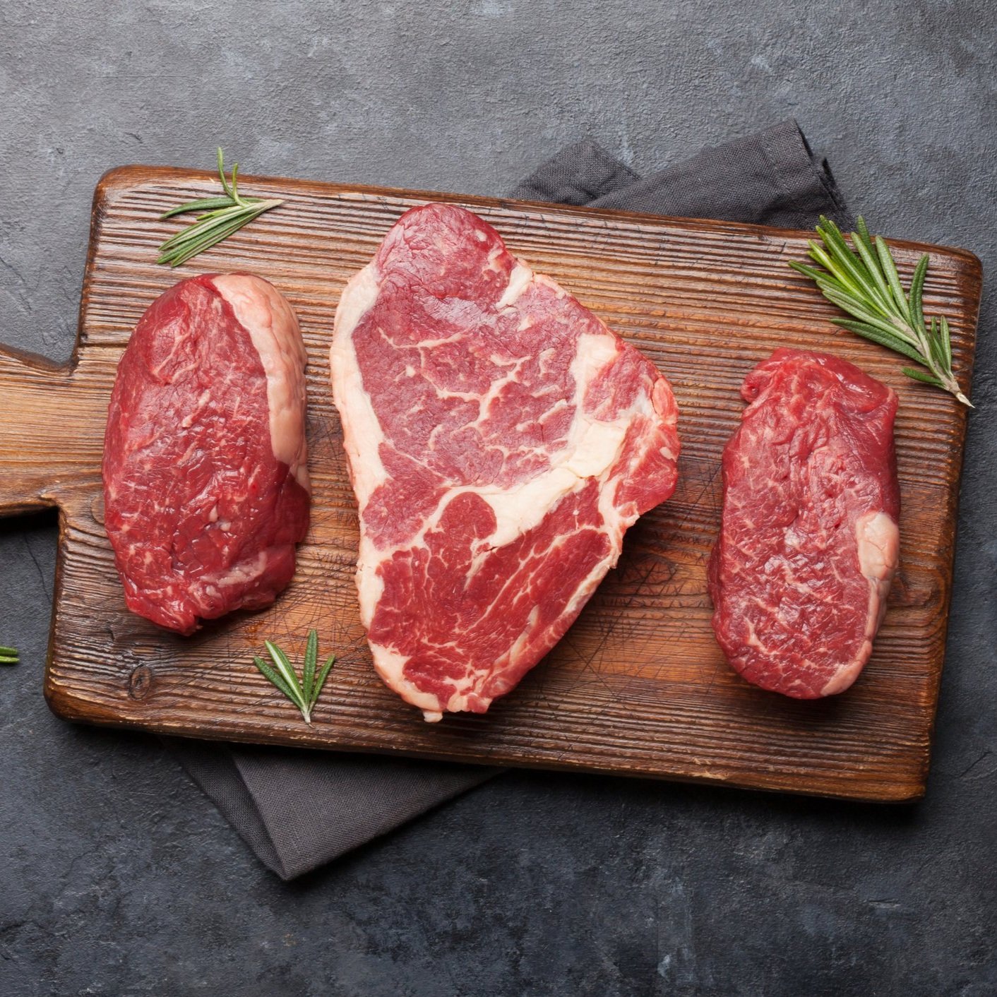 Raw cuts of beef displayed on a wooden cutting board, including ribeye, sirloin, and tenderloin, garnished with fresh rosemary leaves. Ideal for steak recipes and grilling enthusiasts.