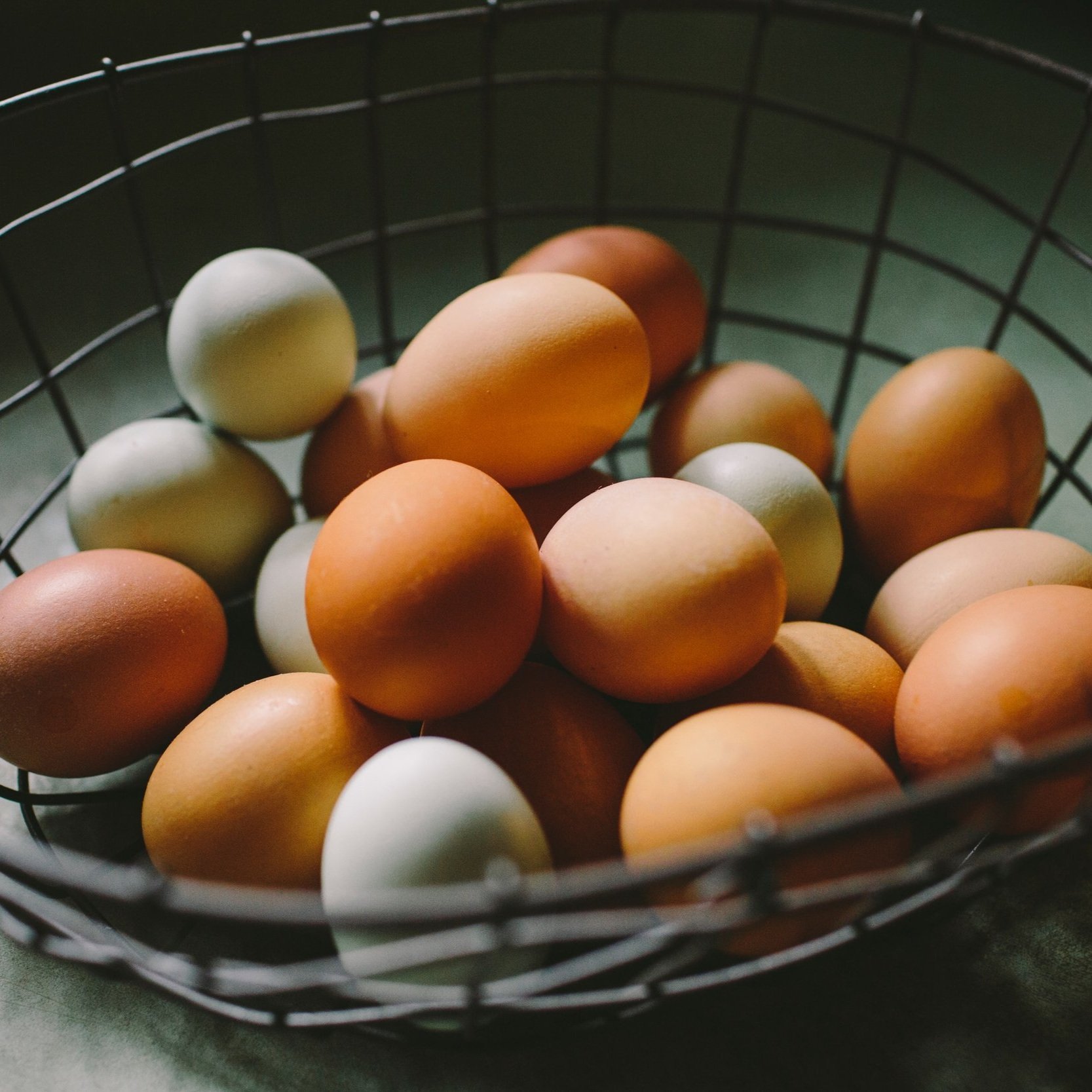 A wire basket filled with various eggs, including brown, tan, and pale blue eggs, arranged on a textured surface.