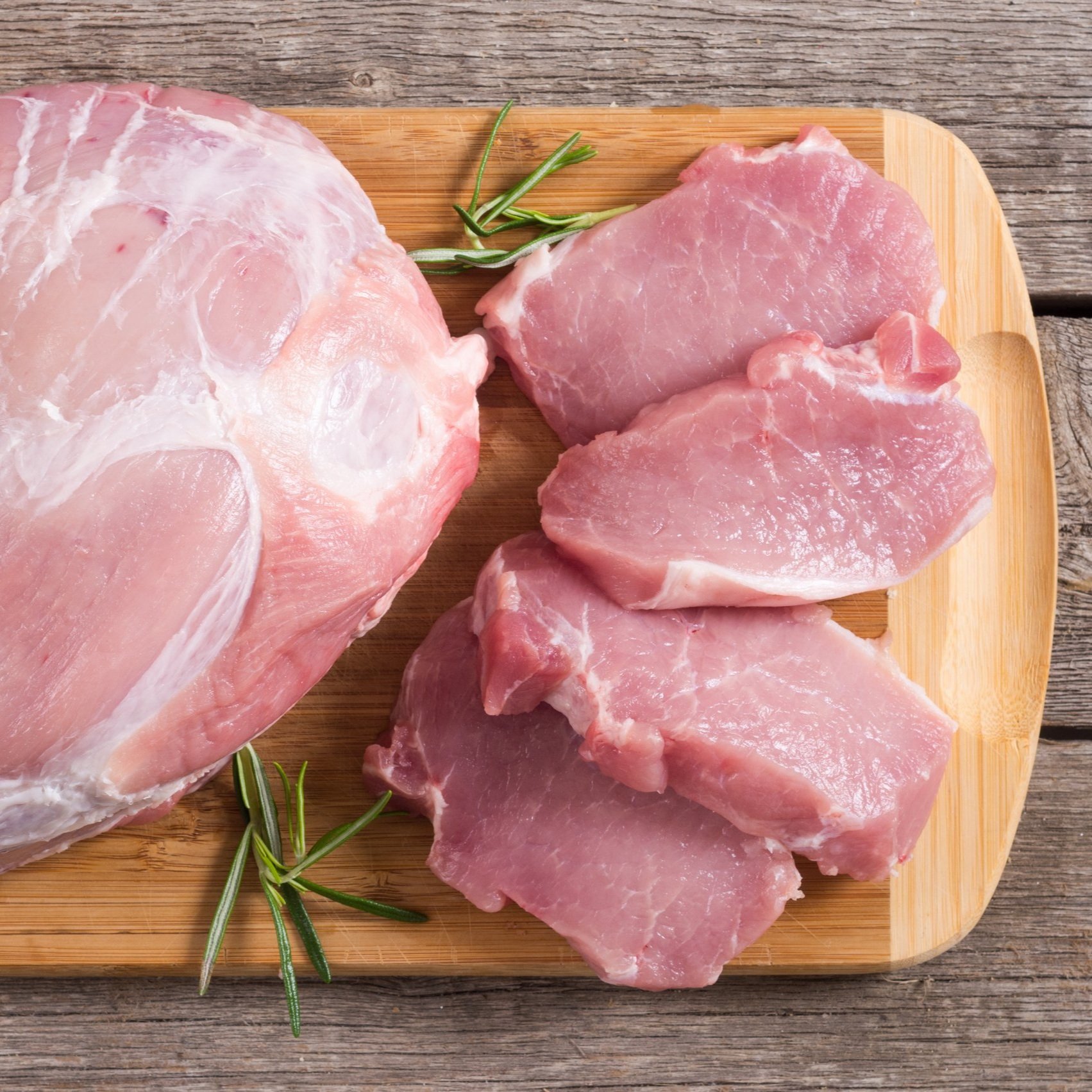 Fresh pork cuts displayed on a wooden cutting board, featuring a large pork shoulder and several neatly sliced pork chops, garnished with sprigs of rosemary.